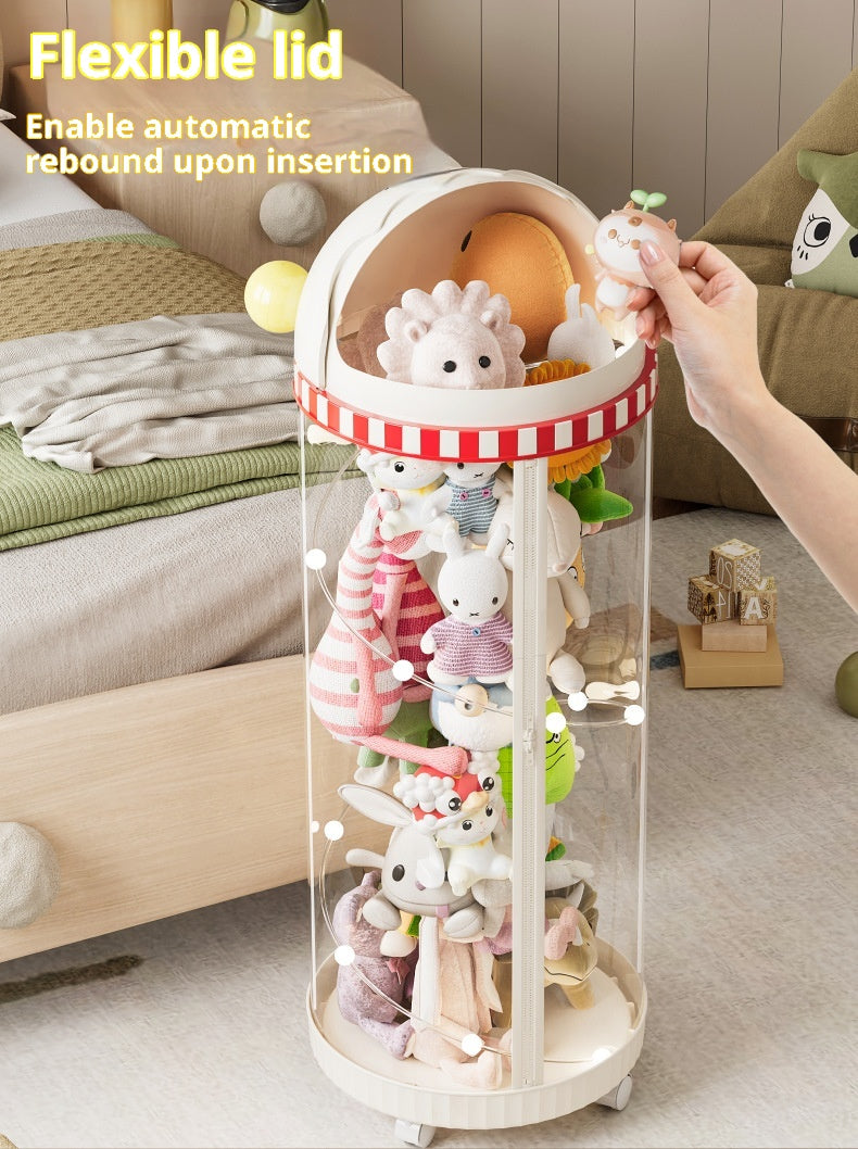 A clear cylindrical toy storage organizer with a cream-colored lid and base, filled with plush animal toys. A human hand is shown inserting a plush toy into the flexible lid, which has text overlay explaining its automatic rebound feature.