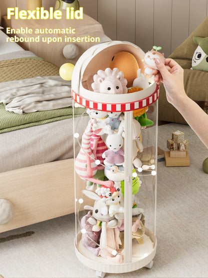 A clear cylindrical toy storage organizer with a cream-colored lid and base, filled with plush animal toys. A human hand is shown inserting a plush toy into the flexible lid, which has text overlay explaining its automatic rebound feature.