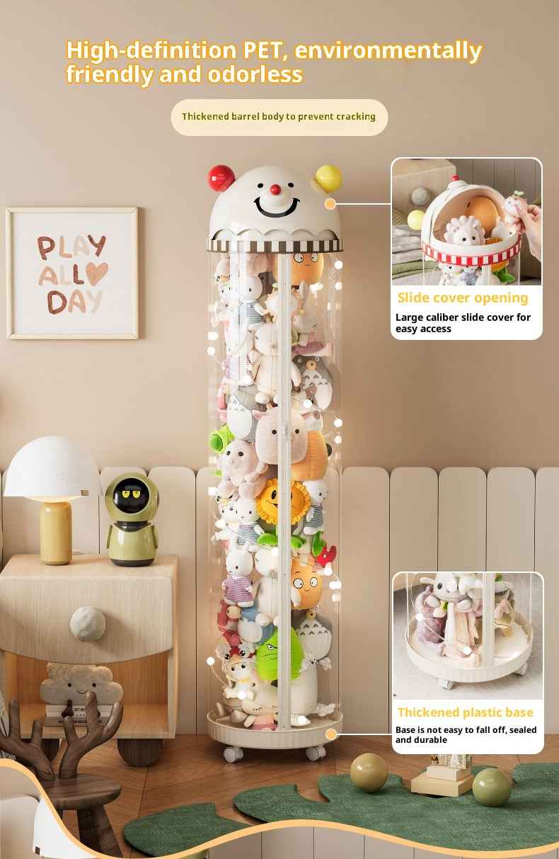 A tall cylindrical collectible display cabinet filled with plush toys, shown in a styled room setting with furniture and decor. The cabinet has a white top and base, a clear barrel body, and warm string lights inside.