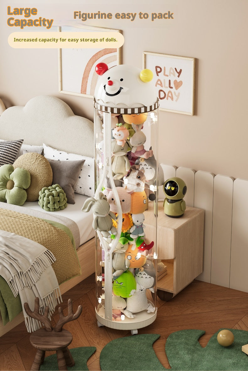 A tall cylindrical clear storage unit filled with plush figurines, shown in a styled bedroom setting. The unit has a white top cap with a smiling face and small colored accents, and warm string lights inside.