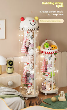 Two decorative hanging string light display stands filled with plush toys in a softly lit bedroom or playroom. The larger left stand is a tall clear cylinder with a white smiling character head on top and warm fairy lights draped inside.