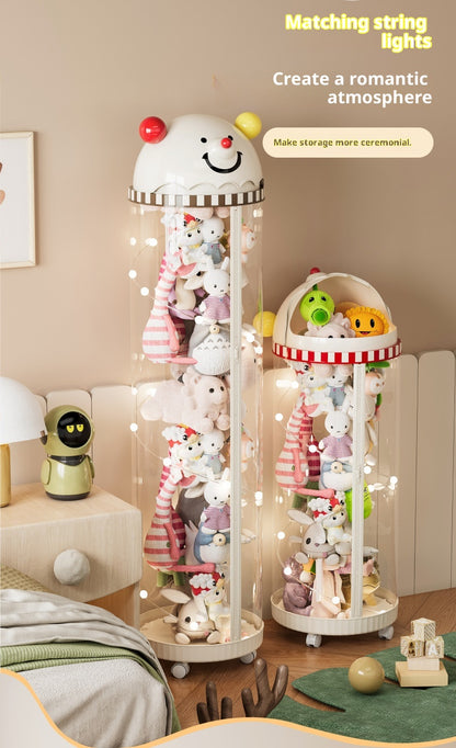Two decorative hanging string light display stands filled with plush toys in a softly lit bedroom or playroom. The larger left stand is a tall clear cylinder with a white smiling character head on top and warm fairy lights draped inside.