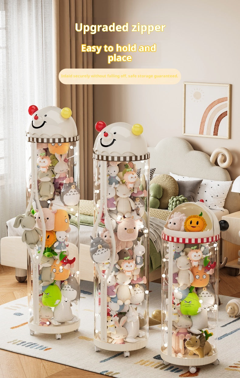 Three clear cylindrical storage bins filled with plush toys, shown in a styled living room setting. The bins have white lids with cartoon face details and a striped top band.
