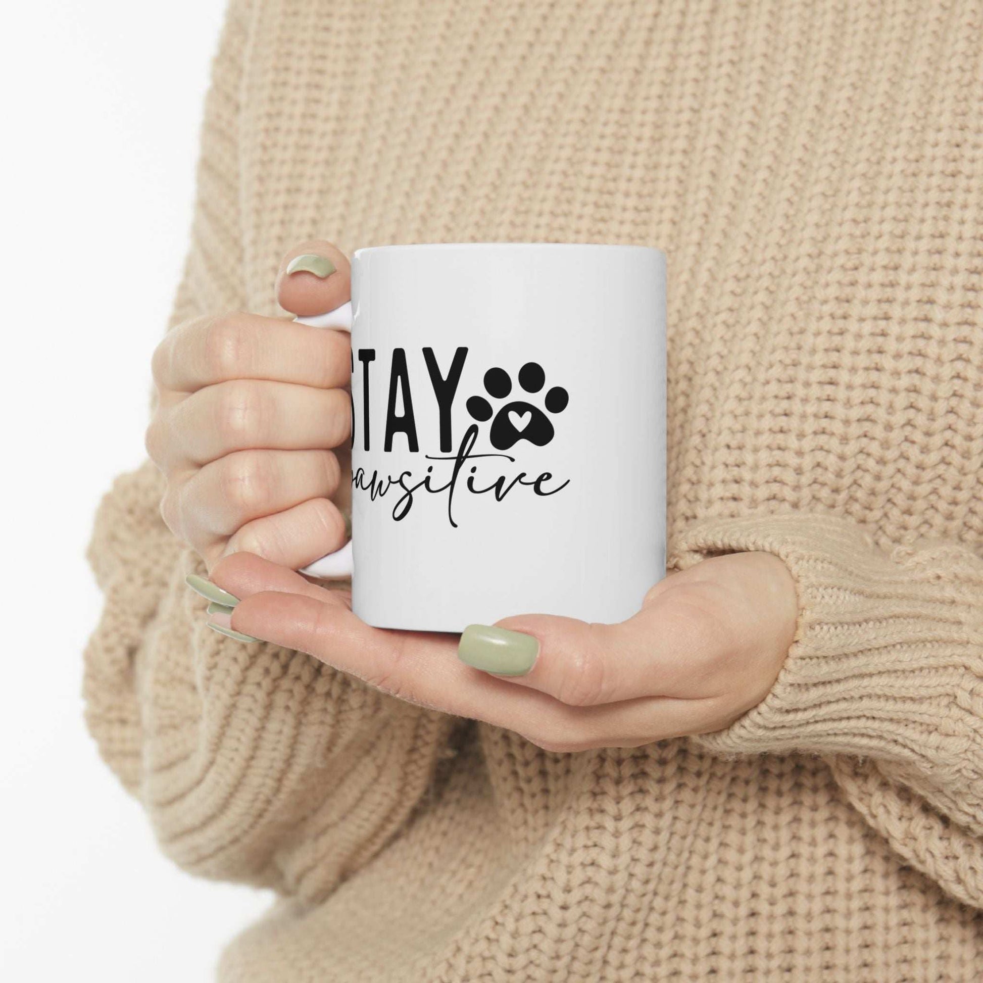 White ceramic Stay Pawsitive Mug held by person wearing beige sweater with green nails